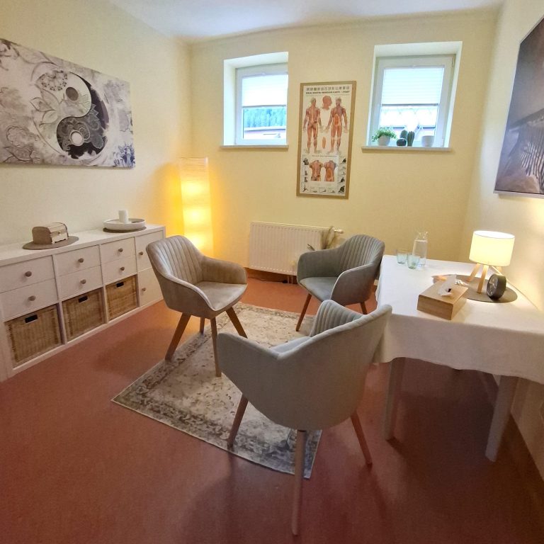 Helles Therapiezimmer mit zwei Sesseln, Tisch und Grafiken an der Wand. Sehr einladend und warm. 